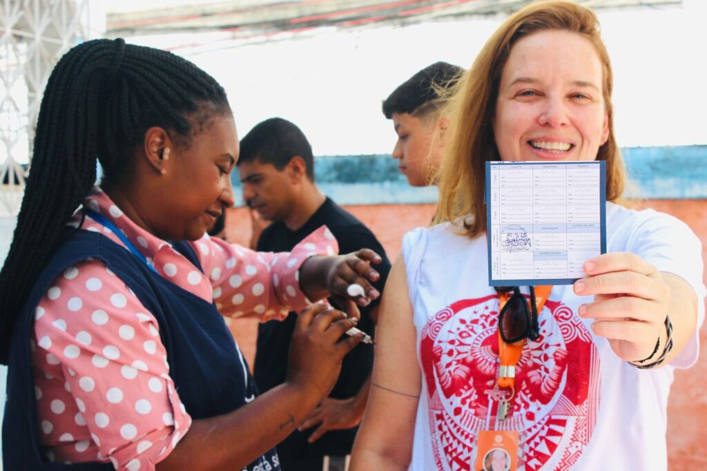 Uma mulher negra com colete do SUS está aplicado uma vacina no braço direito de uma mulher branca, voluntária do Círculo Laranja. Esta mulher está mostrando a carteira de vacinação.