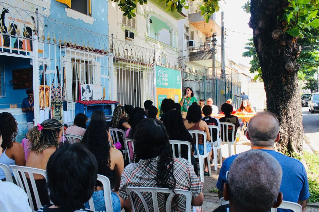 A presidente do Círculo Laranja, Luiette Ornellas, está falando ao microfone e, atrás delas, na mesa de debate, um homem e duas mulheres, nossos três debatedores estão sentados. Há uma plateia de cerca de 20 pessoas sentadas em cadeiras colocadas na calçada. 